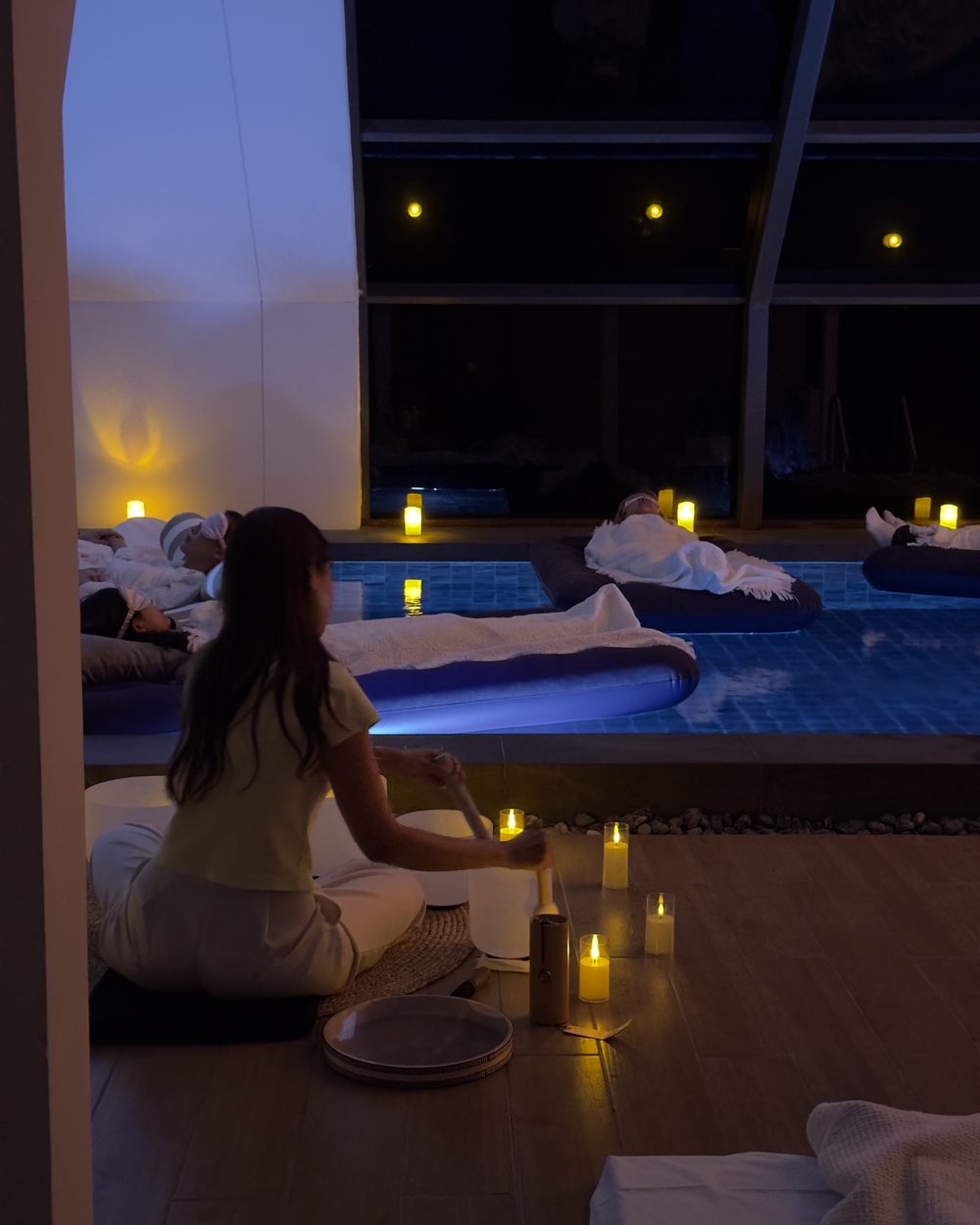 A facilitator playing crystal singing bowls at the pool edge while guests rest on floating beds by candlelight