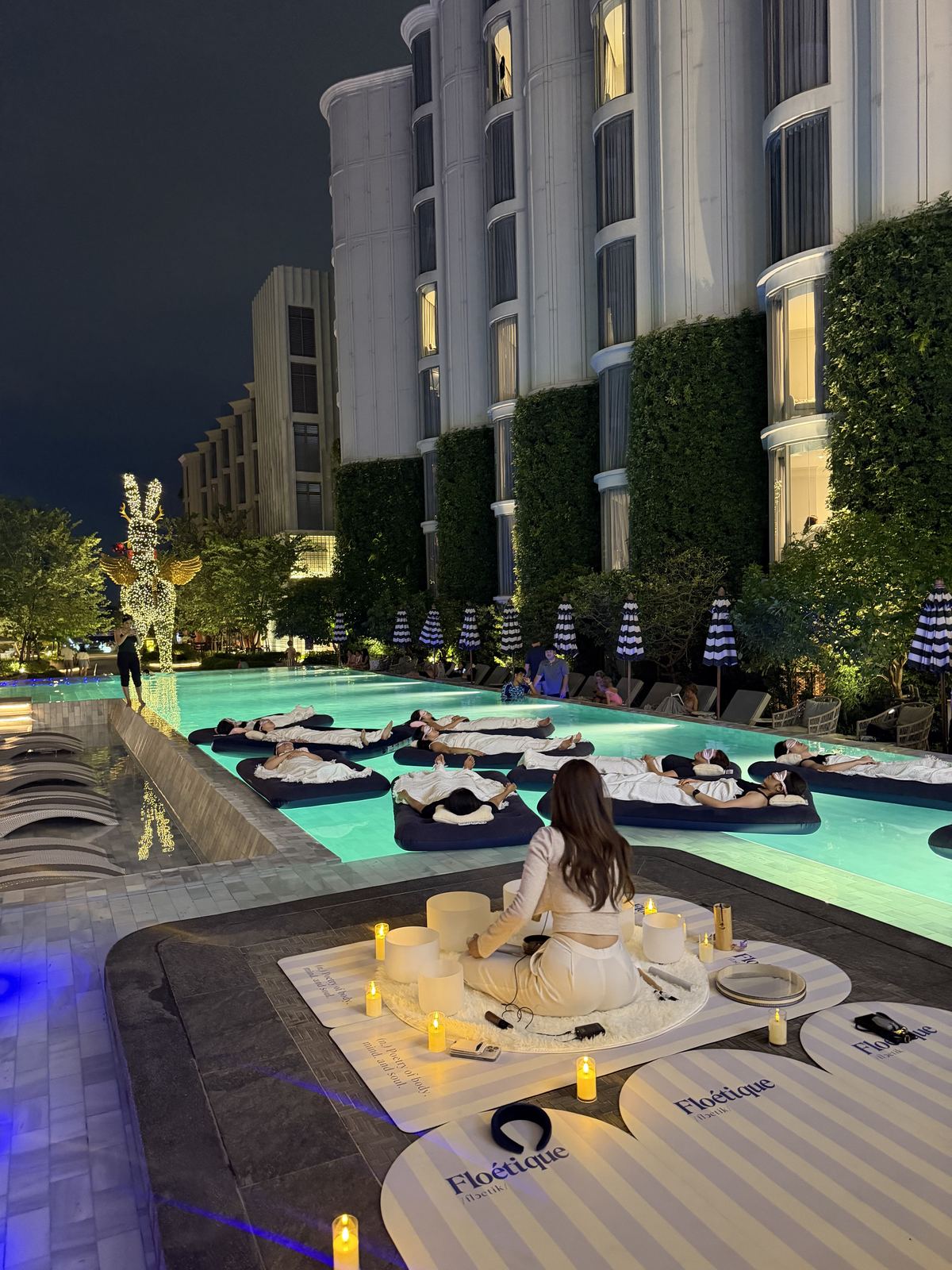 A facilitator with candles and singing bowls beside a hotel pool set with floating beds for a floating sound bath