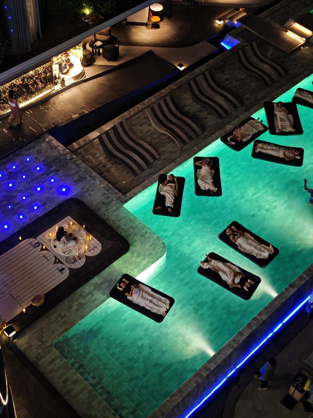 An aerial view of a hotel pool with floating beds and candlelight during a floating sound bath package