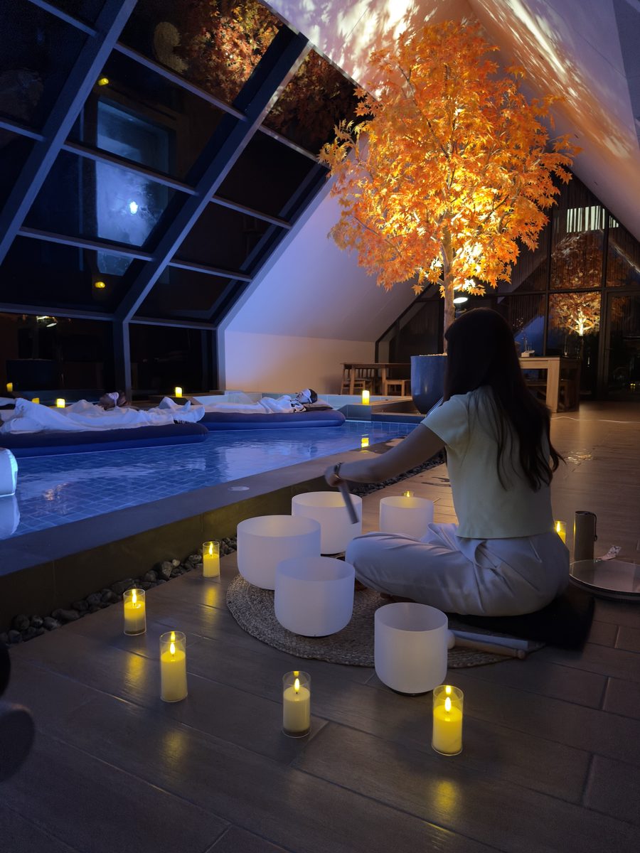 A facilitator playing crystal singing bowls beside a candlelit indoor pool while guests rest on floating beds